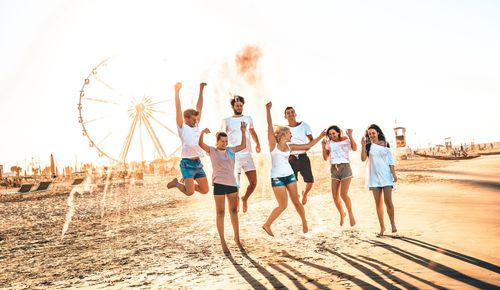 Happy friends group having fun at ferris wheel seaside - Summer vacations friendship concept with millenial guys and girls cheering at public beach - Warm sunset color tone with contrast filtered look